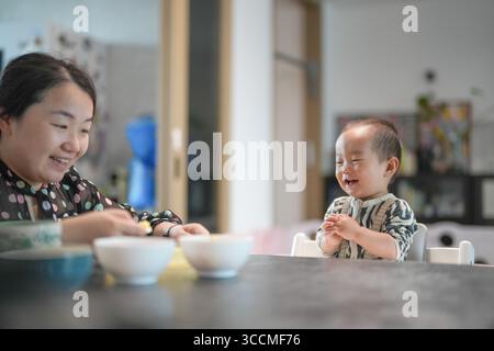 Chinesische Familie im Wohnzimmer sitzt ein einjähriges Baby glücklich in einem Hochstuhl, während ihre Mutter in ihren Dreißigern sie füttert, umgeben von einem gemütlichen Furnit Stockfoto