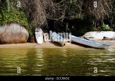 Handwerkliche Fischerboote auf dem Sand eines ruhigen Strandes, mit ruhigem Wasser und üppiger einheimischer Vegetation im Hintergrund. Stockfoto