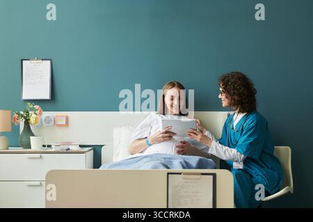 Kaukasische junge Erwachsene Frau sitzt im Krankenhausbett lächelnd, während sie ein digitales Tablet mit einer kaukasischen Schwester mittleren Alters hält, die neben ihr hilft und interagiert Stockfoto