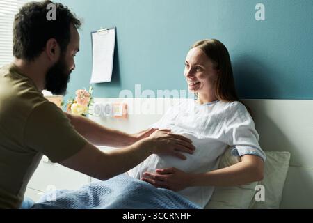 Kaukasische schwangere junge Erwachsene Frau liegend im Krankenhausbett lächelnd, während kaukasischer junger erwachsener Mann ihren Bauch berührte, beide interagierten in der Mutterschaftsmedizin Stockfoto