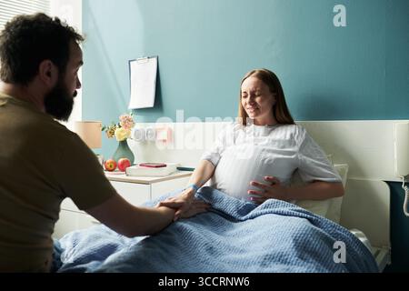 Kaukasische schwangere junge Erwachsene Frau, die Wehenschmerzen erlebt, die im Krankenhausbett liegt und Magen hält, während kaukasischer bärtiger junger erwachsener Mann sie mit der Hand unterstützt Stockfoto