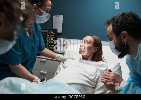 Kaukasische junge Erwachsene Frau, die im Krankenhausbett liegt umgeben von zwei weiblichen Ärzten und kaukasischen jungen erwachsenen Mann, medizinisches Personal, das den Patienten während der Geburt unterstützt Stockfoto