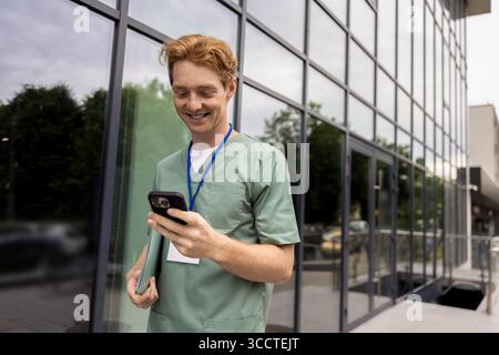 Rothaariger Arzt, der während der Pause in der Nähe eines reflektierenden Glasgebäudes auf dem Handy surft Stockfoto