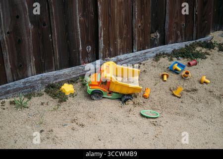 Bunte Sandkistenspielzeuge aus Kunststoff, die in trockenem Sand in der Nähe eines Holzzauns verstreut sind. Kinderspiel, Outdoor-Lernen, frühe Entwicklung, unstrukturierte Erkundung Stockfoto