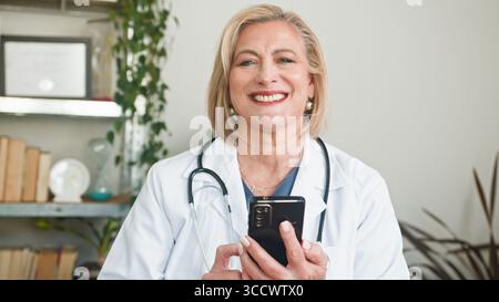 Lächelnder Seniorarzt hält Smartphone im professionellen Klinikbüro Stockfoto