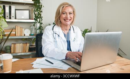 Lächelnder Oberarzt, der an einem Laptop in einem modernen Büro arbeitet Stockfoto