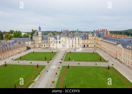 Aus der Vogelperspektive auf Château de Fontainebleau, wo der braune Stein des Palastes einen Kontrast zu den lebhaften grünen Rasenflächen bildet, eingerahmt vom grauen Himmel, Fontainebleau, Île-de-France, Frankreich. Stockfoto