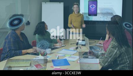 Präsentation einer Frau im mittleren Erwachsenenalter mit Gesten am Konferenztisch im Büro, mit Laptops und Projektionsleinwand Stockfoto