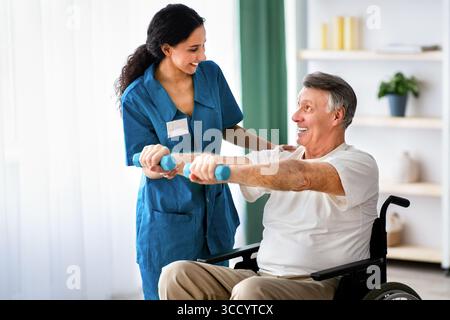 Physiotherapeutin hilft älteren Mann im Rollstuhl Übungen mit Hanteln im Gesundheitszentrum Stockfoto