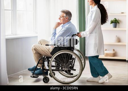 Seitenansicht eines jungen Arztes, der älteren behinderten Männern im Rollstuhl hilft, drinnen Stockfoto