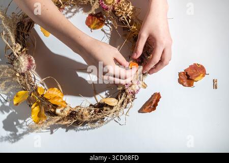 Frauenhände kreieren einen Herbstkranz aus natürlichen Materialien auf weißem Hintergrund. Thanksgiving-Tag-Konzept. Obere V Stockfoto