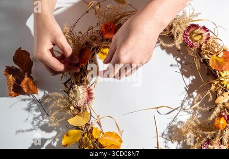 Frauenhände kreieren einen Herbstkranz aus natürlichen Materialien auf weißem Hintergrund. Thanksgiving-Tag-Konzept. Obere V Stockfoto