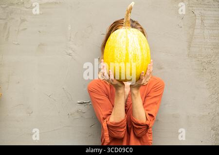 Eine gesichtslose Frau hält einen großen gelben Kürbis in den Händen vor dem Hintergrund einer Zementwand. Kopierbereich. Thanksgiving Day Konzept Stockfoto