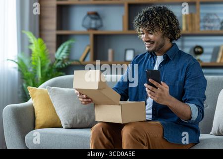 Ein fröhlicher junger Mann, der auf einer Couch sitzt und einen Karton öffnet, während er sein Smartphone hält. Die Umgebung ist ein modernes Wohnzimmer mit einem komfortablen Stockfoto