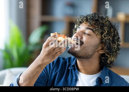 Ein junger Mann mit lockigen Haaren genießt ein Stück Pizza und genießt den Geschmack in einer gemütlichen Wohnumgebung mit verschwommenem Hintergrund für Ruhe Stockfoto