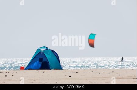 Einsames Zelt an einem Sandstrand im Sommer, mit einem verschwommenen Kiteboarder, der im Hintergrund surft, mit der glitzernden Oberfläche des sonnendurchfluteten Ozeans Stockfoto