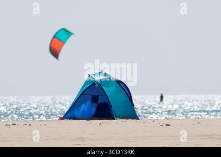 Einsames Zelt an einem Sandstrand im Sommer, mit einem verschwommenen Kiteboarder, der im Hintergrund surft, mit der glitzernden Oberfläche des sonnendurchfluteten Ozeans Stockfoto