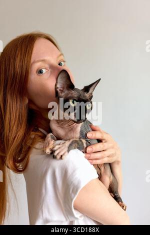Eine rothaarige Frau, die ein weißes Hemd trägt, hält ihre sphynx-Katze liebevoll vor einem neutralen Hintergrund. Die auffällige Verbindung zwischen dem Haustier und dem Besitzer ist ev Stockfoto