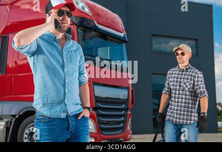 Zwei Männer unterhalten sich vor einem Transportunternehmen vor einem roten Truck und diskutieren tagsüber Logistik. Stockfoto