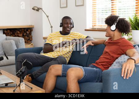 Verschiedene männliche Freunde, die Podcasts im Wohnzimmer auf blauem Sofa mit Kopfhörern und Mikrofon aufnehmen Stockfoto
