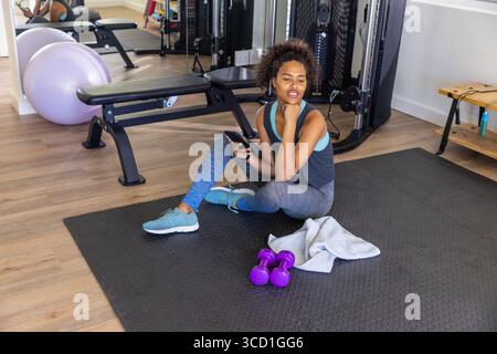 Afroamerikanerin, die auf der Matte mit Kurzhanteln ruht, Smartphone im Fitnessstudio überprüft, Kopierraum Stockfoto