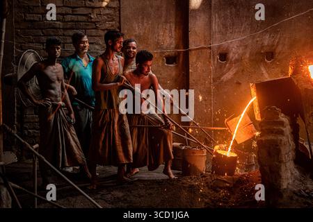 Bogura, Bangladesch - 07. Oktober 2021: Blick auf intensive Hitze und geschmolzenes Metall, das die Gesichter von Arbeitern in einer Gießerei beleuchtet, deren Silhouetten stark gegen das feurige Leuchten sind. Stockfoto