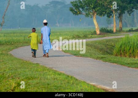 Bogura, Bangladesch - 09. Oktober 2020: Blick auf zwei Figuren, die auf einem gewundenen Pfad durch üppige grüne Felder unter dem sanften Morgenlicht spazieren und eine ruhige und friedliche ländliche Szene schaffen. Stockfoto