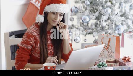 Eine lächelnde junge Frau, die auf ihrem Laptop im Internet surft, um Weihnachten zu verhandeln, während sie an einem Tisch vor dem geschmückten Baum in ihrer Re sitzt Stockfoto