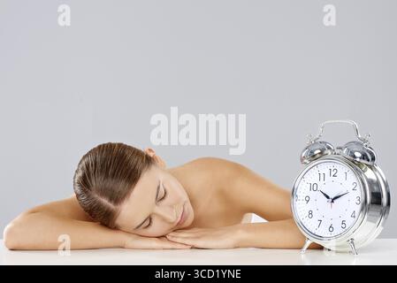 Schöne junge Frau, die neben einem klassischen Retro-Wecker schläft, mit Glocken, die an einem Tisch sitzen, mit dem Kopf auf dem Arm und einem friedlichen Aufenthalt Stockfoto