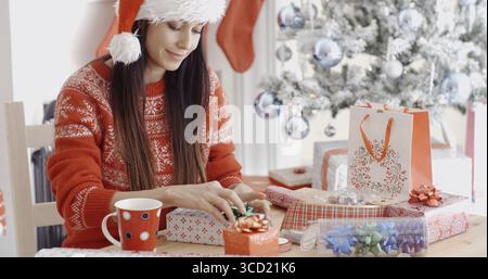 Junge Frau, die ihre Weihnachtsgeschenke mit Schleifen und Bändern dekoriert, während sie an einem Tisch vor einem geschmückten Weihnachtsbaum sitzt Stockfoto