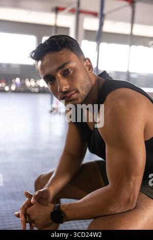 Muskulöser Mann, der auf dem Boden des Fitnessstudios sitzt und sich nach vorne gelehnt hat und die Smartwatch in der Nähe des Pull-up-Rigs überprüft Stockfoto