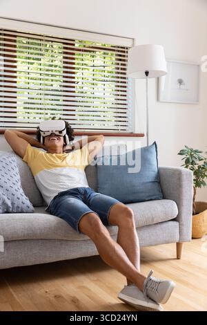 Afroamerikanischer Mann, der sich im Wohnzimmer auf einem grauen Sofa liegend und ein Virtual-Reality-Headset trug Stockfoto