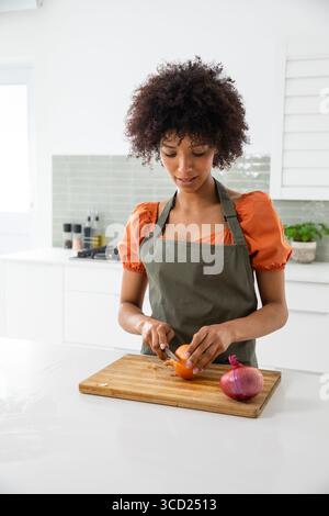 Afroamerikanerin in grüner Schürze, die gelbe Zwiebel mit Messer auf Schneidebrett in der Küche schneidet Stockfoto