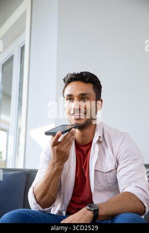 Mann, der auf dem Sofa im Wohnzimmer sitzt und in das Smartphone spricht, mit sichtbarer Armbanduhr Stockfoto