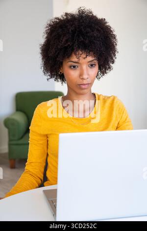 Afroamerikanische Frau, die an silbernem Laptop am weißen Schreibtisch im Heimbüro mit grünem Sessel arbeitet Stockfoto