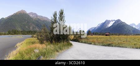 Blick auf einen Schotterpfad, der sich durch goldene Wiesen schlängelt und zu schneebedeckten Bergen unter klarem Himmel führt, eine ruhige Landschaft Alaskas, Anchorage, Alaska, USA. Stockfoto