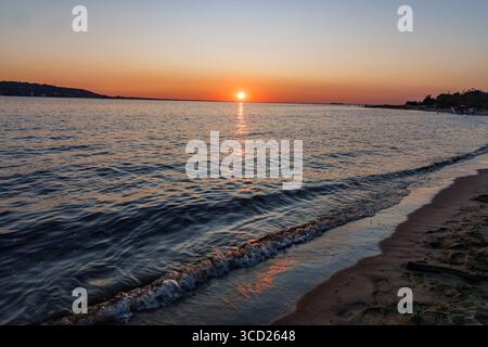 Friedlicher Sonnenuntergang über ruhigen Meereswellen, die sich auf den Sandstrand mit orangefarbenem Himmel und entfernter Küste im ruhigen Abendlicht ergießen. Stockfoto