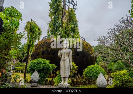 Stehende Buddha-Statue vor der Kuppel der Hölle im Xieng Khuan Buddha Park in Vientiane, Laos, umgeben von üppiger tropischer Vegetation Stockfoto