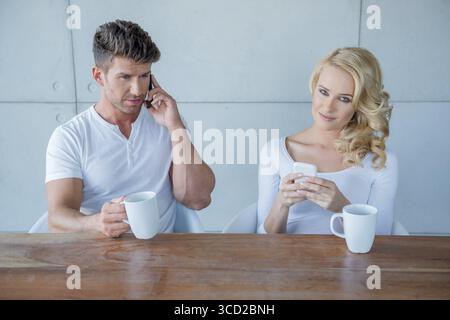 Weißes sitzendes Paar in lässiger Kleidung, das Kaffee am Holztisch trinkt, während es mit seinem eigenen Handy beschäftigt ist Stockfoto