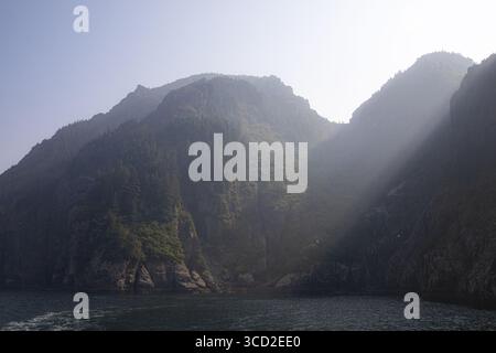Aus dem dunklen Wasser der Resurrection Bay erhebt sich der Blick auf dramatische Klippen, die von einem mystischen Dunst des Sonnenlichts umgeben sind, Kenai Stockfoto