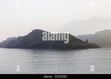 Blick auf eine zerklüftete Insel mit dunklem, felsigem Gelände und karger Vegetation liegt im ruhigen Wasser der Resurrection Bay unter den nebeligen Bergen der Kenai Fjorde Stockfoto