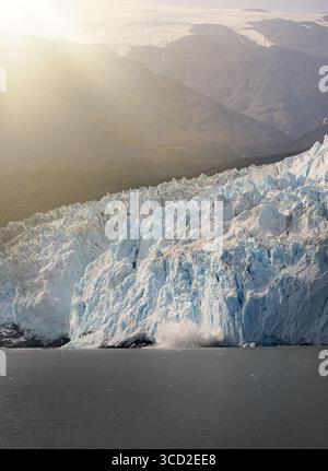 Der Blick auf das eisblaue Gesicht eines Gletschers trifft dramatisch auf das dunkle Wasser der Aialik Bay unter einem sanften, goldenen Sonnenlicht, Berge im Hintergrund von K Stockfoto