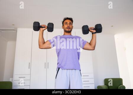 Asiatischer Mann, der schwarze Hexen-Kurzhanteln auf Schulterhöhe in der Heimtrainer in lila Sportswear hebt Stockfoto