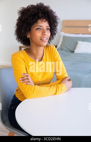 Afroamerikanerin sitzt am Tisch im Schlafzimmer und trägt senfgelbe Oberseite und schaut nach rechts Stockfoto