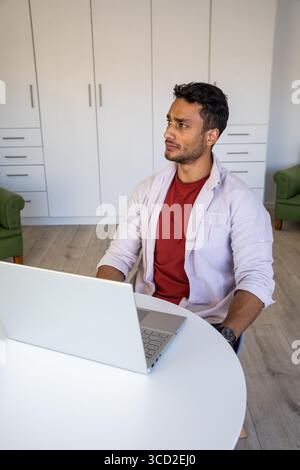 Mann sitzt im Home Office an einem weißen runden Tisch mit Laptop in der Nähe von Schränken, Kopierraum Stockfoto