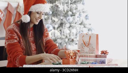 Junge Frau, die ihre Weihnachtsgeschenke mit Schleifen und Bändern dekoriert, während sie an einem Tisch vor einem geschmückten Weihnachtsbaum sitzt Stockfoto
