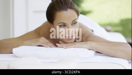 Die neugierige Frau auf dem Spa-Tisch, die in weiße Handtücher gewickelt ist, schaut zur Seite, während sie ihr Kinn auf ihren Händen legt Stockfoto