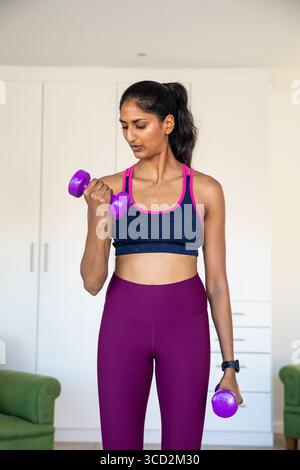 Indische Frau in Sportswear hält lila Kurzhanteln und arbeitet im Wohnzimmer Stockfoto