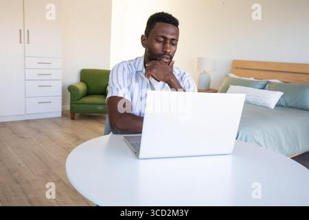 Afroamerikanischer Mann sitzt am weißen Tisch im hellen Schlafzimmer und arbeitet am Laptop neben dem Bett Stockfoto