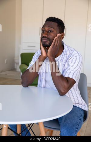 Afroamerikaner sitzt an einem runden weißen Tisch und blickt nach Hause in der Nähe eines grünen Sessels Stockfoto